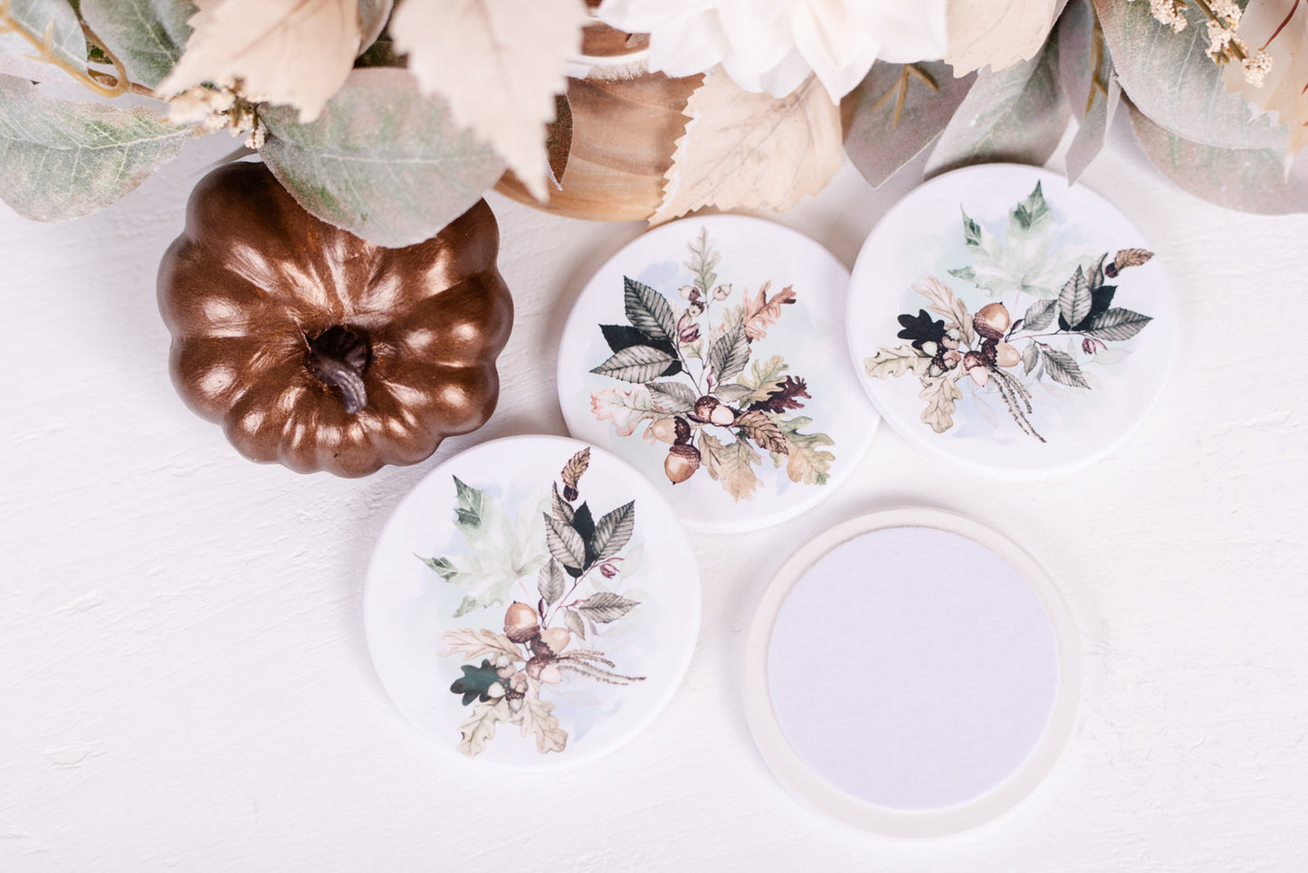 Neutral coloured fall, ceramic coasters with felt backing on a white table with pumpkins and foliage
