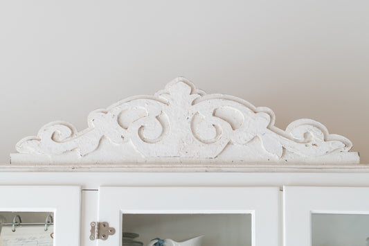 Decorative white pediment molding above a white cabinet with glass doors.