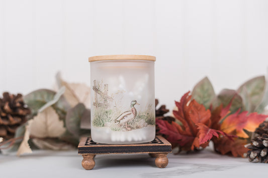 Candle jar with a wooden lid and base, surrounded by autumn leaves and pinecones on a light background