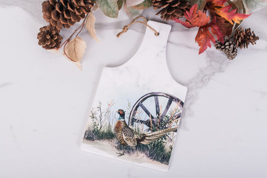 Trivet with a pheasant and wagon wheel design on a marble surface with autumn leaves and pine cones.