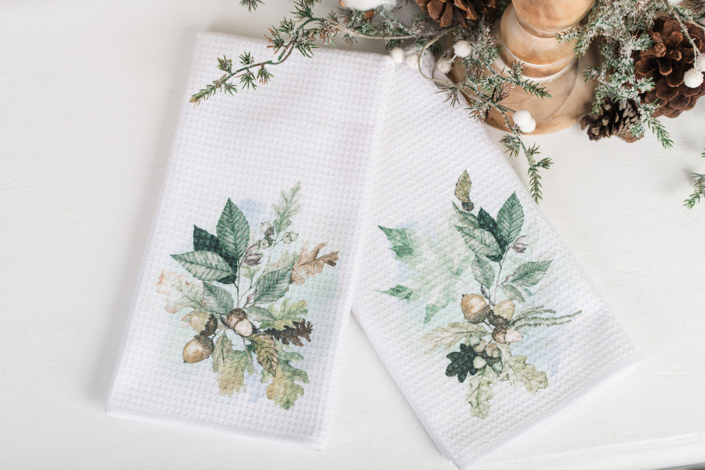 Two white waffle weave tea towels with a green floral design, displayed on a table with a pine branch decor in the background.