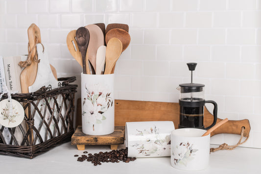 A magnolia utensil holder, styled with wooden spoons, sits on a farmhouse kitchen counter alongside a French press and a coffee canister filled with coffee beans.
