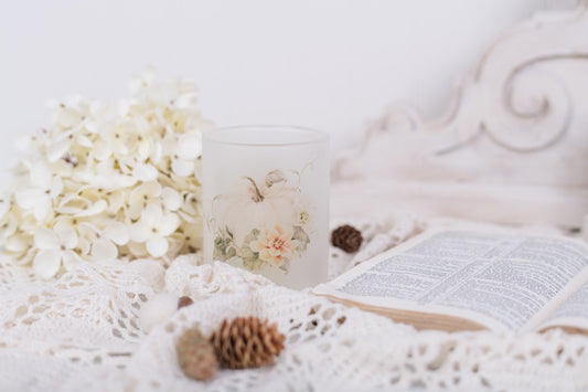 Candle with a pumpkin and floral design next to an open book on a textured surface