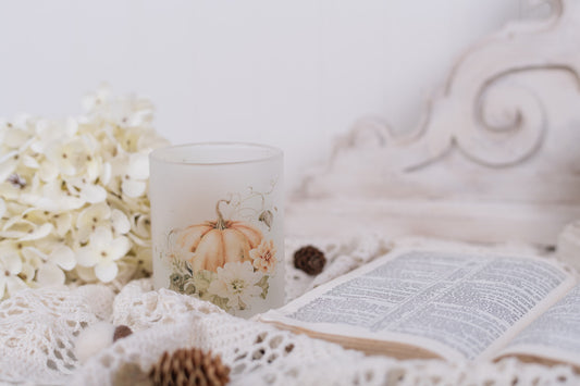 Candle with pumpkin design on a white surface with flowers and an open book.