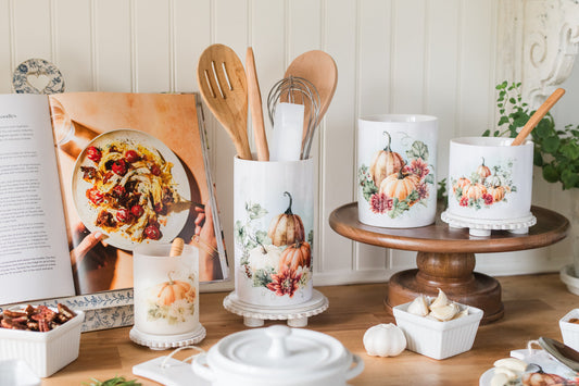 Decorative fall kitchen items including a cookbook, wooden spoons, and ceramic containers with pumpkin designs on a wooden table.
