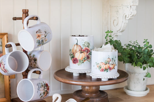Decorative mugs with floral designs on a stand, next to a stand with canisters with similar decorations on a wooden table.