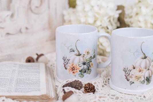 Two white mugs with pumpkin and floral designs on a textured surface with an open book and pinecones.