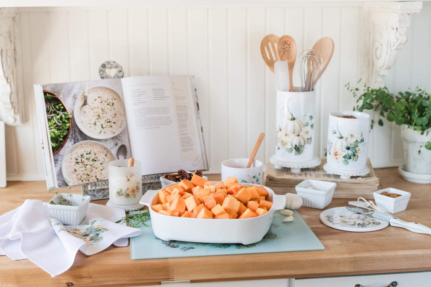 Scripture cutting board styled with kitchen canisters and ingredients, with a casserole dish full of butternut squash sitting on the glass board as a hot plate.