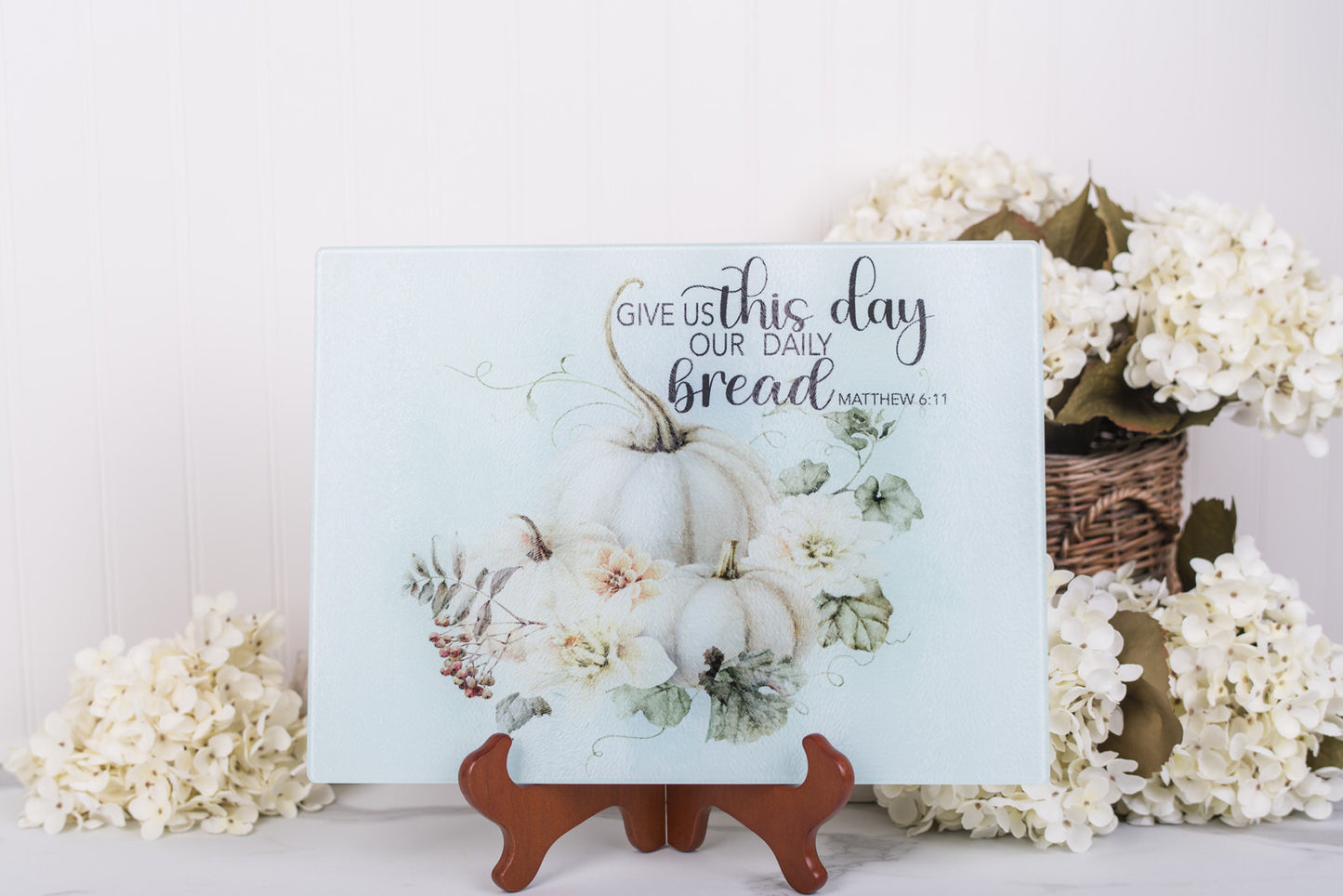 Fall glass cutting board with pumpkins and flowers on a stand, surrounded by white flowers.