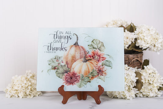 Glass fall cutting board with pumpkins and flowers on a stand, surrounded by white flowers.