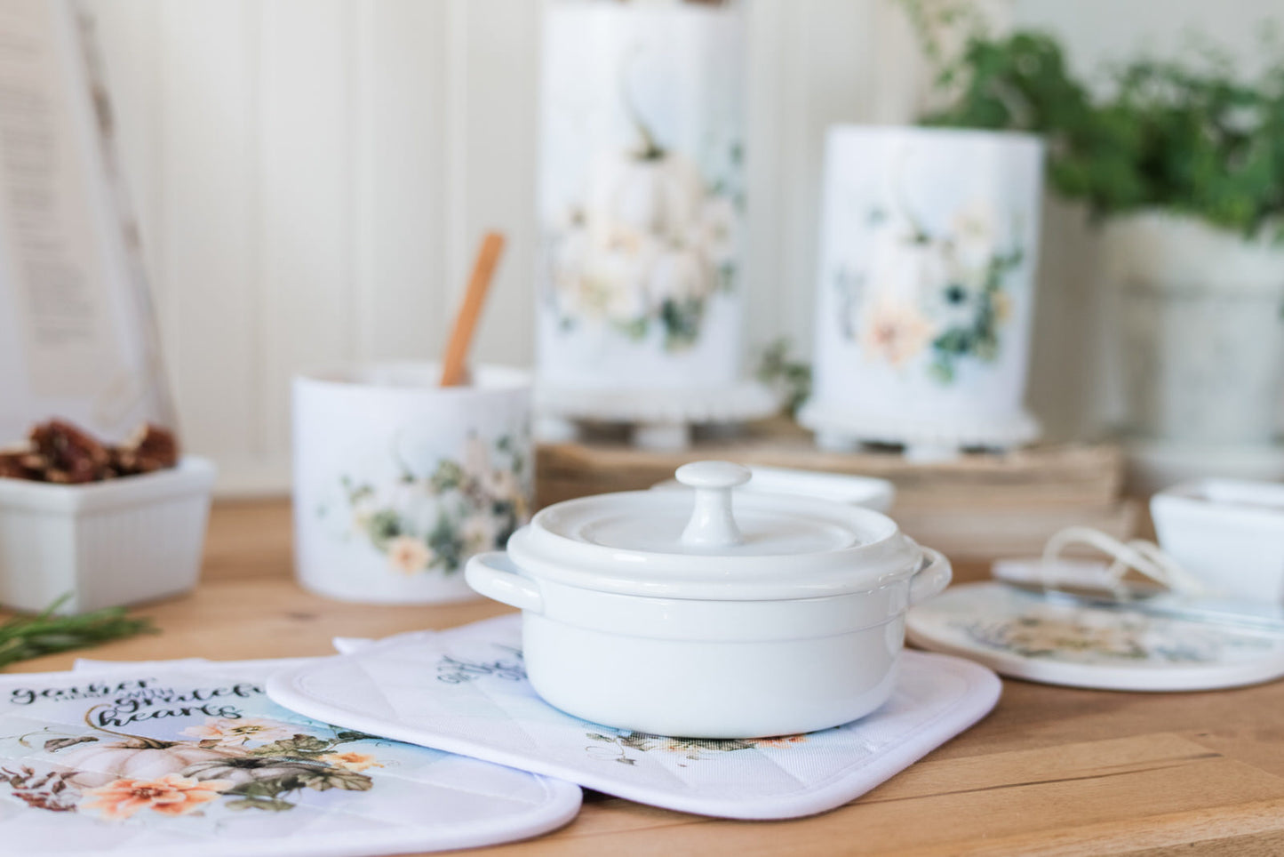 White ceramic pot sitting on fall pot holders, on a wooden table with pumpkins and a floral design and greenery in the background