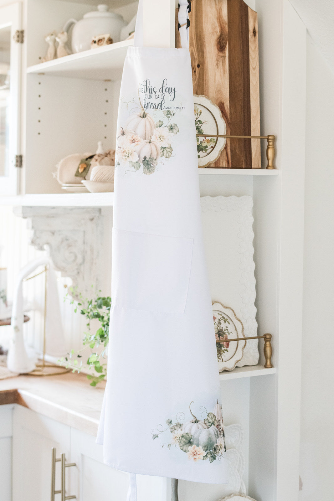 White apron with floral designs hanging on a white shelf in a kitchen.