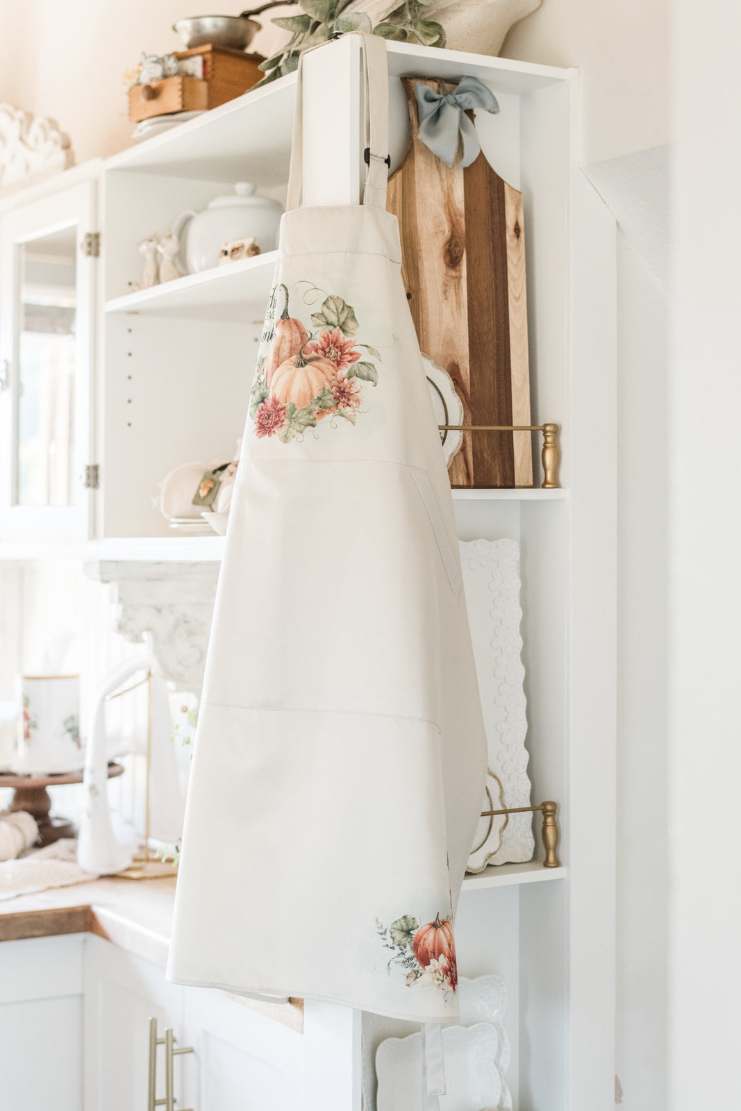 Beige apron with floral design hanging on a white shelf in a kitchen.