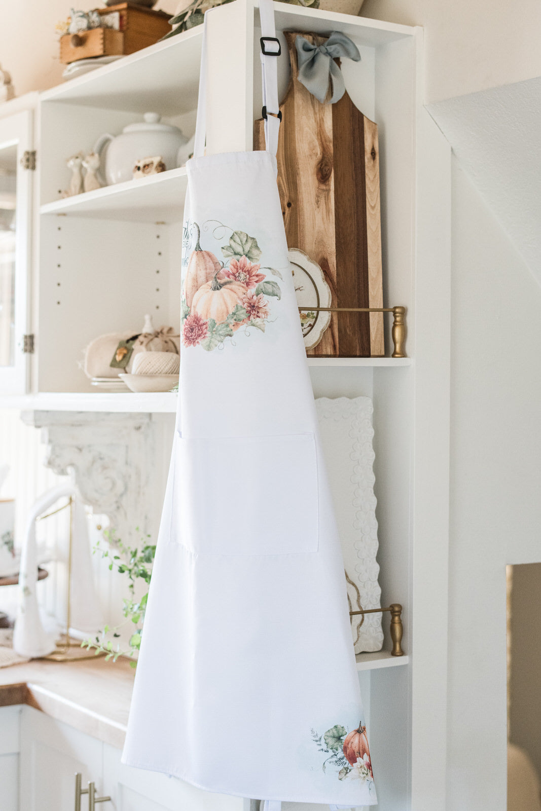 White apron with fall pumpkins and floral design hanging on a white shelf in a kitchen.