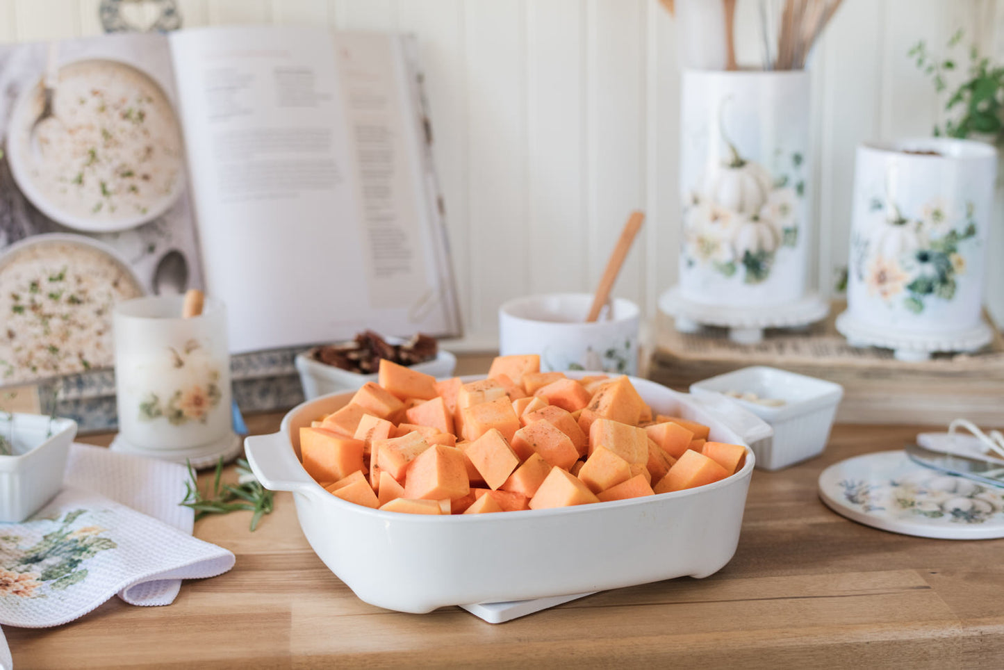 White casserole dish filled with sliced squash on a trivet, protecting wooden countertop with decorative items in the background.