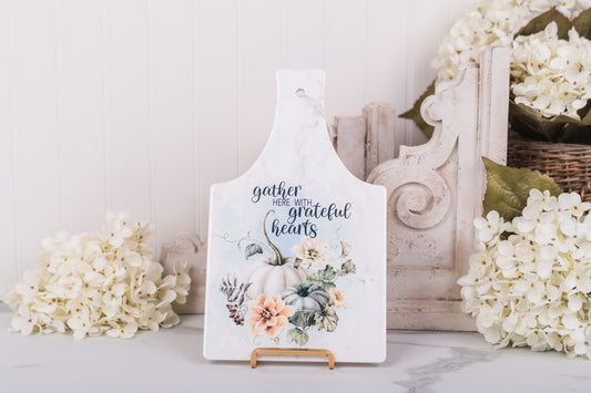 Fall kitchen trivet featuring a floral design and the text 'gather here with grateful hearts' on a marble surface adorned with flowers.
