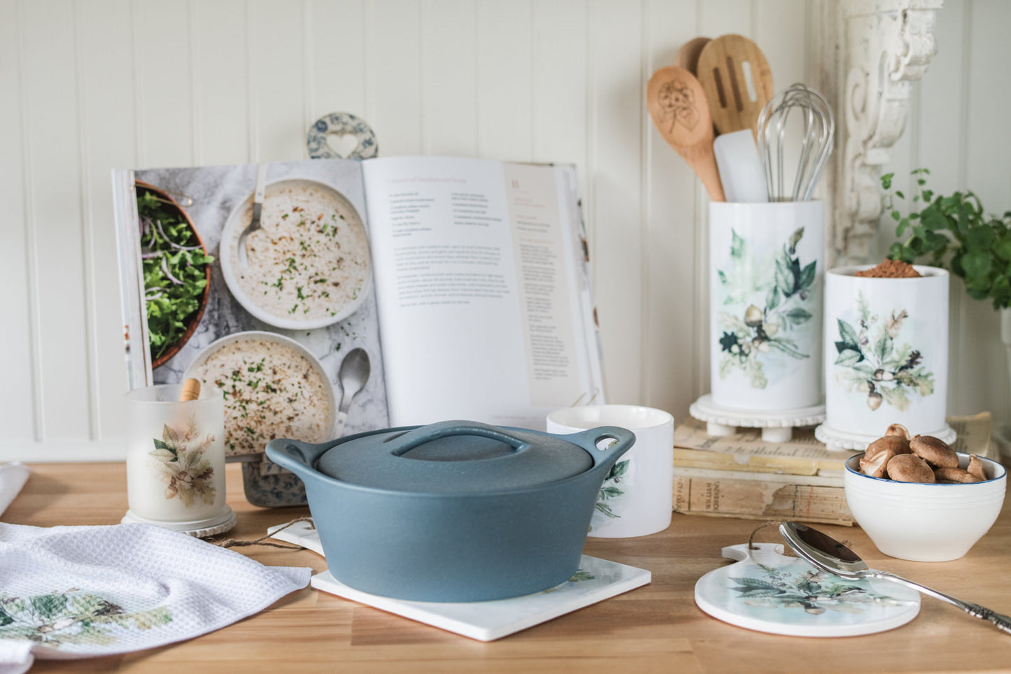Blue cast iron cookware on a wooden table with cookbooks and kitchen utensils in the background. Ceramic utensil holder with watercolour oak leaves and acorns
