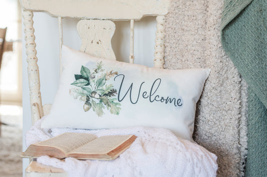 Decorative pillow with 'Welcome' text and floral design on a cozy chair with an open book.