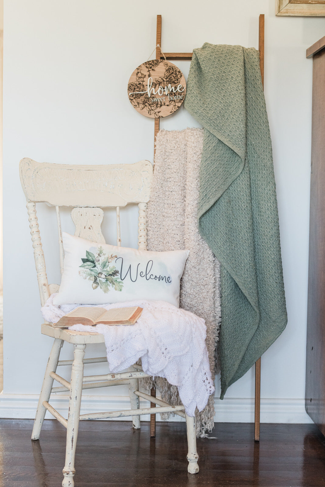 Chair with a pillow and blanket, wooden ladder with green blanket and 'Welcome' sign, against a white wall.