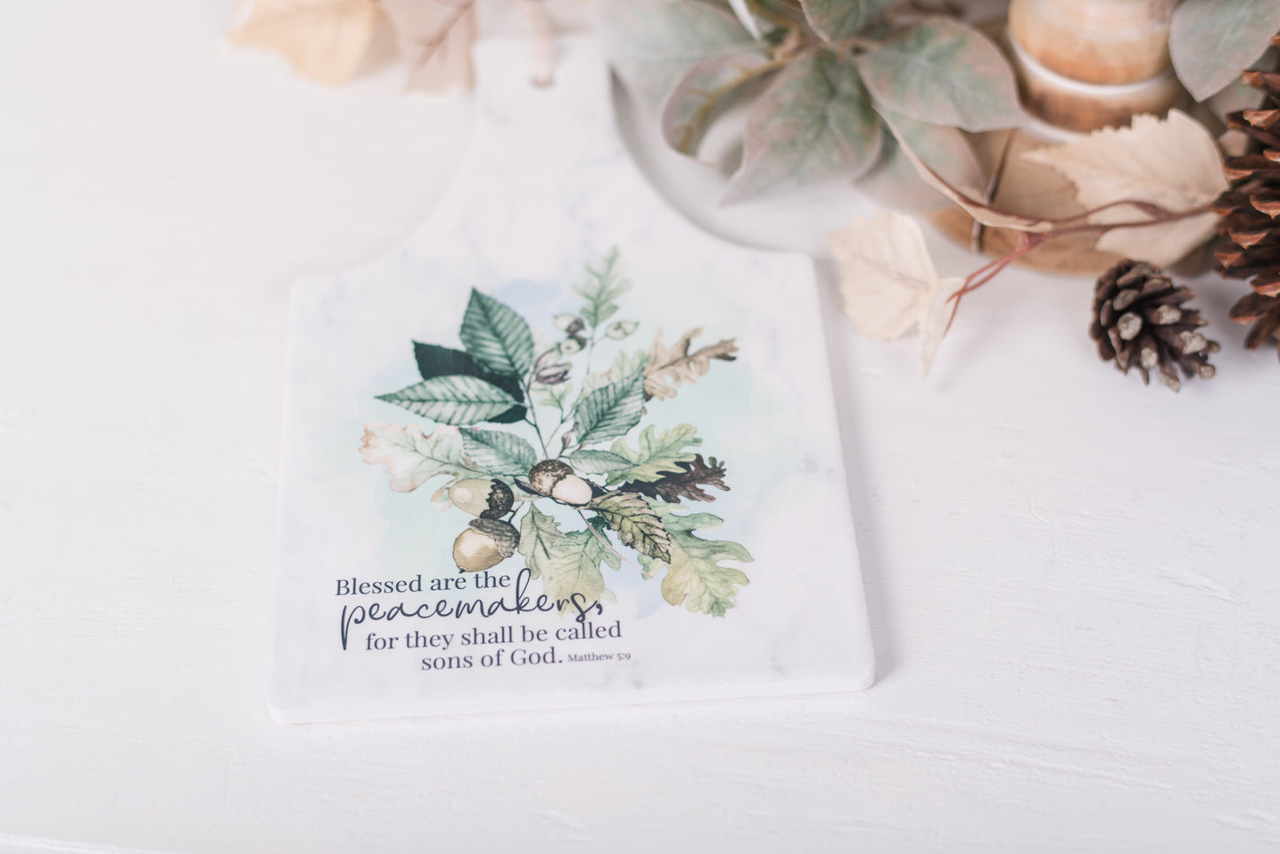 White ceramic trivet with scripture for Matthew 5:9 with fall leaves, and acorns styled on a countertop