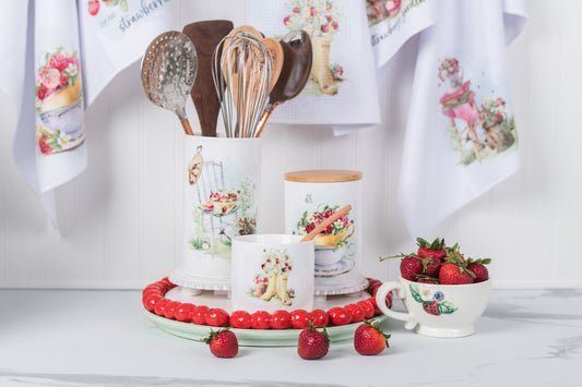 Kitchen-themed decor with cups, spoons, and a bowl of strawberries on a white surface.