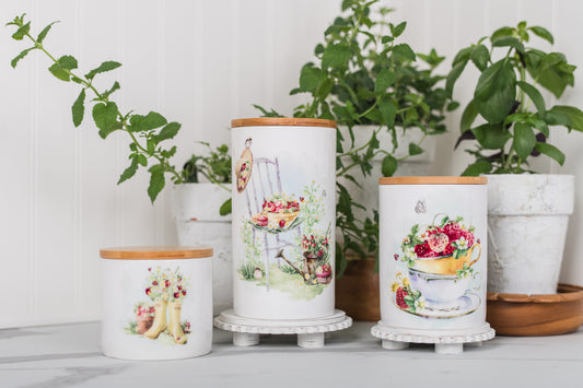 Three decorative canisters with strawberry designs on a surface with plants in the background
