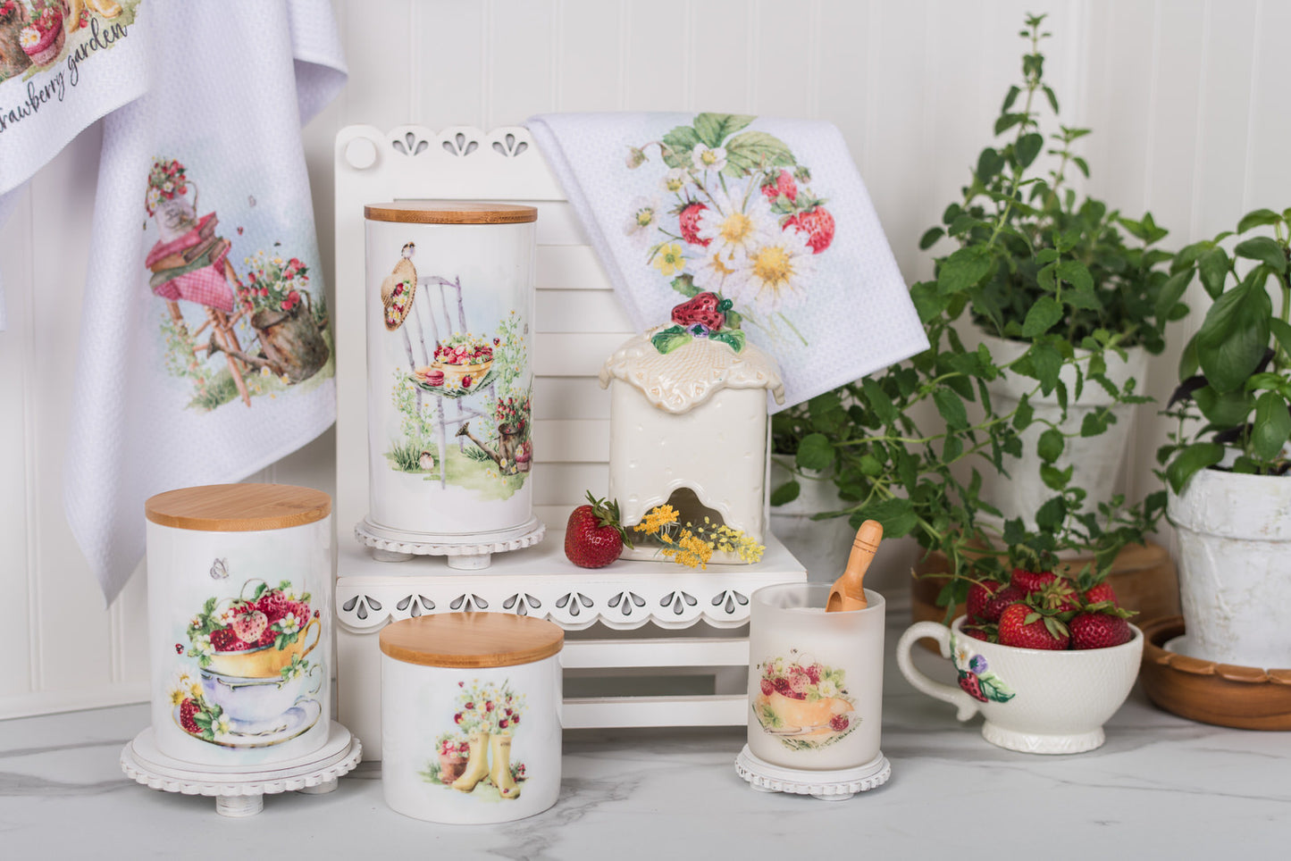 Decorative kitchen canisters with strawberry designs on a white surface with plants in the background.