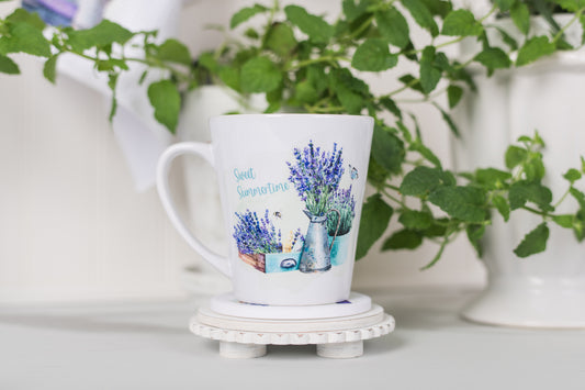 White mug with lavender design on a white surface with green plants in the background