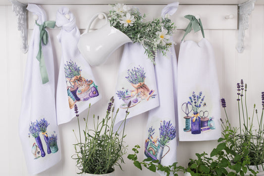 Set of towels with lavender designs hanging on a white wall with plants around.
