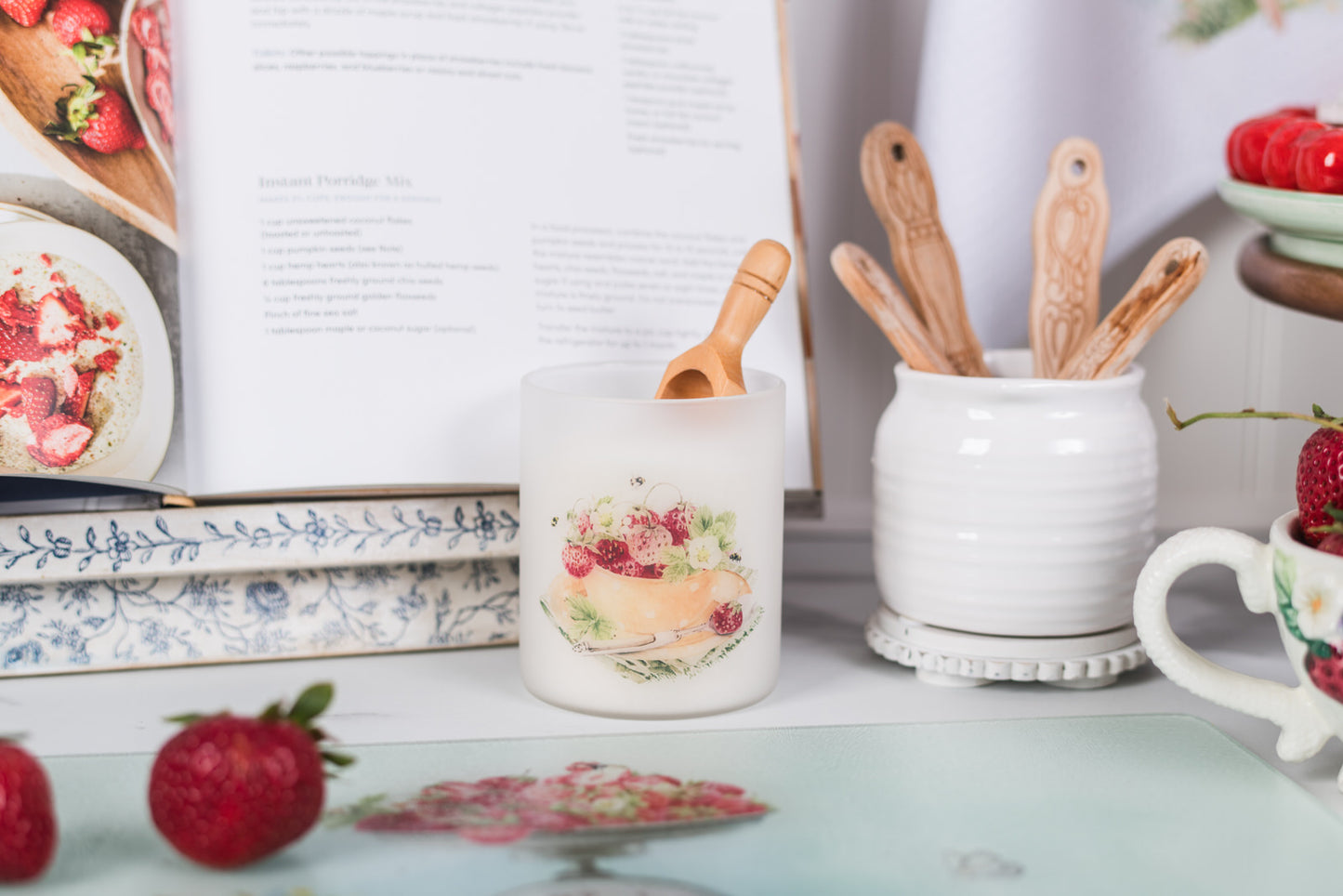 Candle holder with fruit design and wooden spoons in a white container on a decorative surface.
