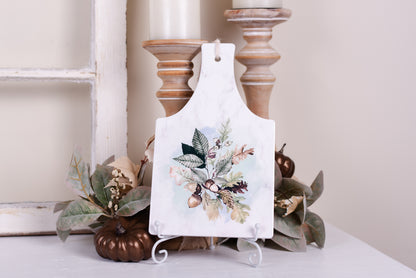 Neutral fall ceramic trivet with acorns and fall leaves, sitting on an easel styled with pumpkins and foliage and candlesticks 