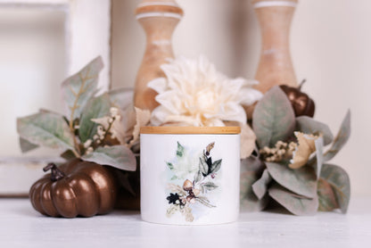 A small ceramic canister with a bamboo lid, featuring an acorn and leaf pattern, is placed on a surface with decorative items around it.