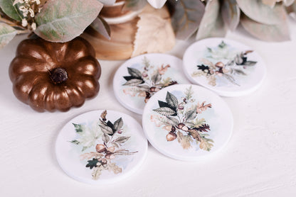 A set of four white ceramic coasters with a floral and woodland foliage pattern, placed on a table with a pumpkin-shaped decoration in the background.