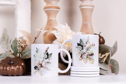 Two white ceramic coffee mugs with a floral and woodland foliage design, placed on a surface with autumn-colored leaves and decorative items in the background.