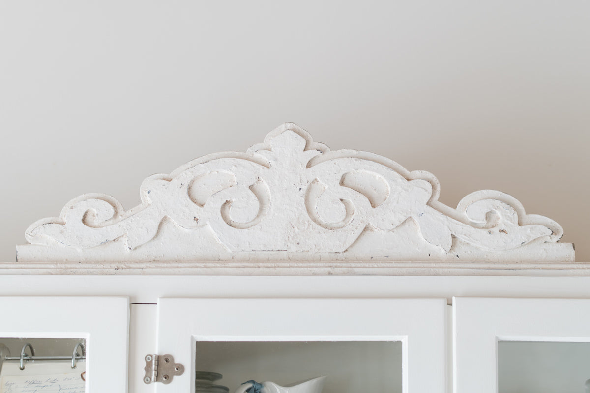 Decorative white pediment molding above a white cabinet with glass doors.