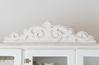 Decorative white pediment molding above a white cabinet with glass doors.