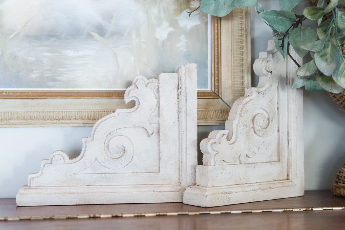Decorative white corbels on a shelf with framed artwork and greenery in the background