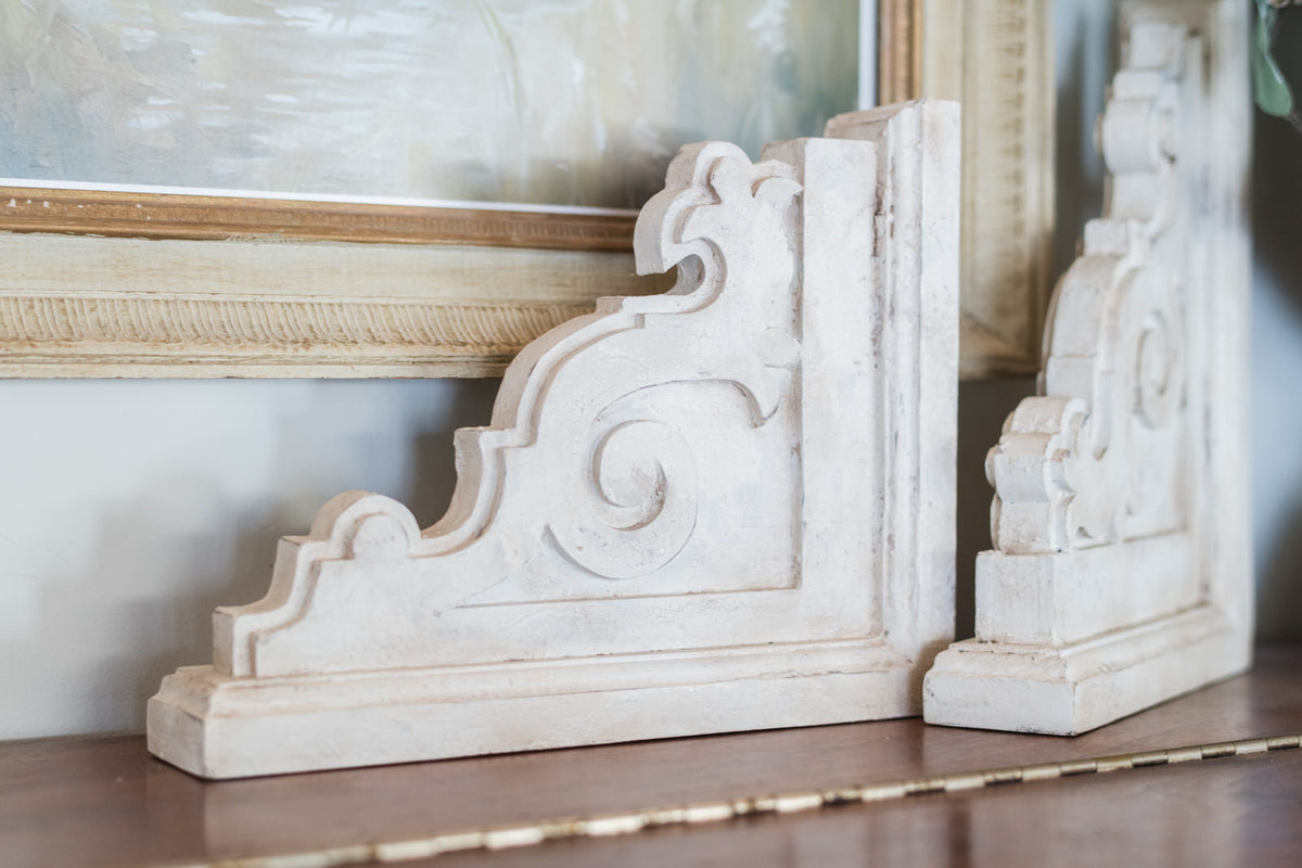 Decorative white corbels on a wooden surface with a blurred painting in the background