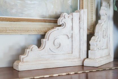 Decorative white corbels on a wooden surface with a blurred painting in the background
