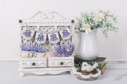 Decorative white cabinet with lavender details, flowers, and a vase on a white surface.