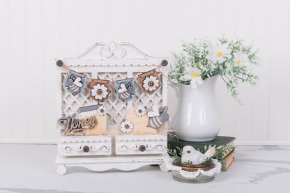 interchangeable cottage cabinet with banner across top with a 'Honey Bee' banner, bird figurine, and vase with flowers on a white surface.
