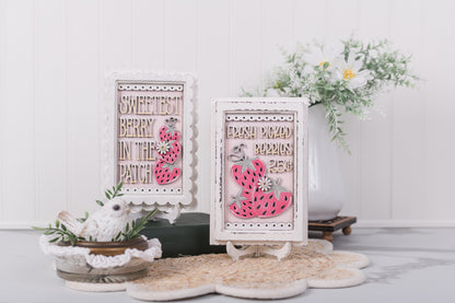 Decorative DIY signs with strawberry designs and text on a white surface with plants.