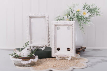 Two white picture frames on a decorative stand with a small bird figurine and floral arrangement in the background.