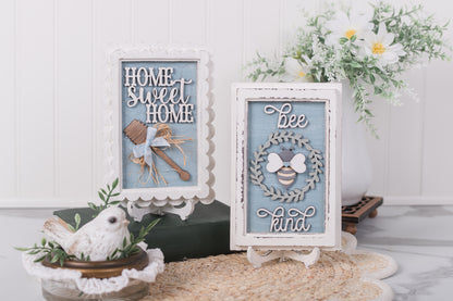 Decorative signs with 'Home Sweet Home' and 'Be kind' text, surrounded by plants and a white bird figurine.