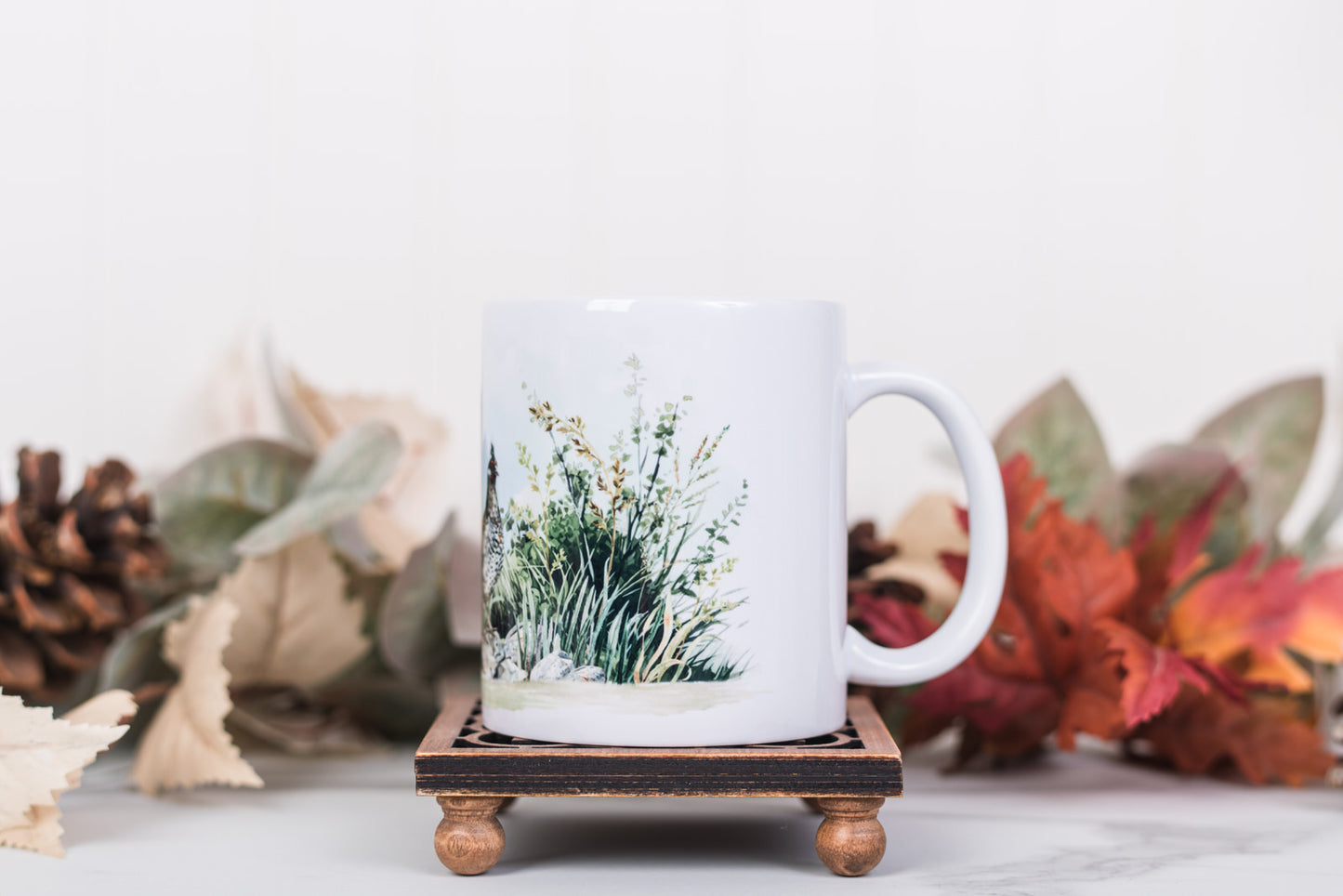 White mug with nature-themed design on a wooden coaster, surrounded by autumn leaves and pinecones.