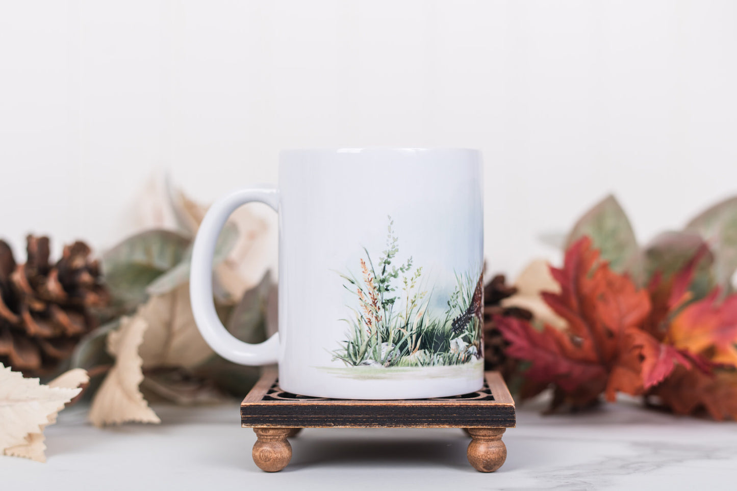 White mug with nature-themed design on a wooden coaster with fall leaves and pinecones in the background.