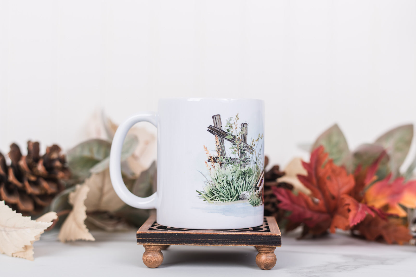 White mug with nature-themed design on a wooden stand with autumn leaves and pinecones in the background