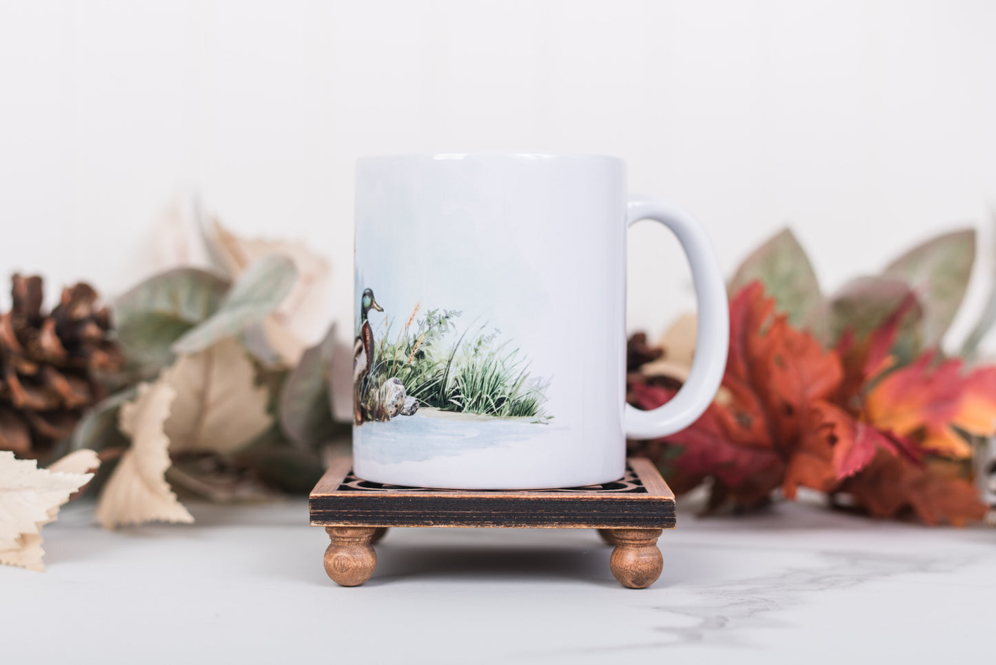 White mug with a nature-themed design on a wooden stand, surrounded by autumn leaves and pine cones.