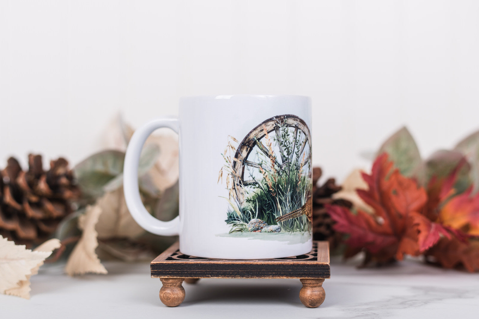 White mug with nature-themed design on a wooden coaster, surrounded by autumn leaves and pinecones.