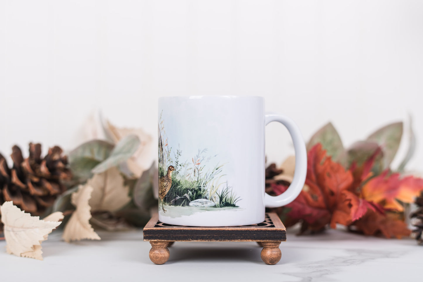 White mug with nature-themed design on a wooden stand with fall decorations in the background